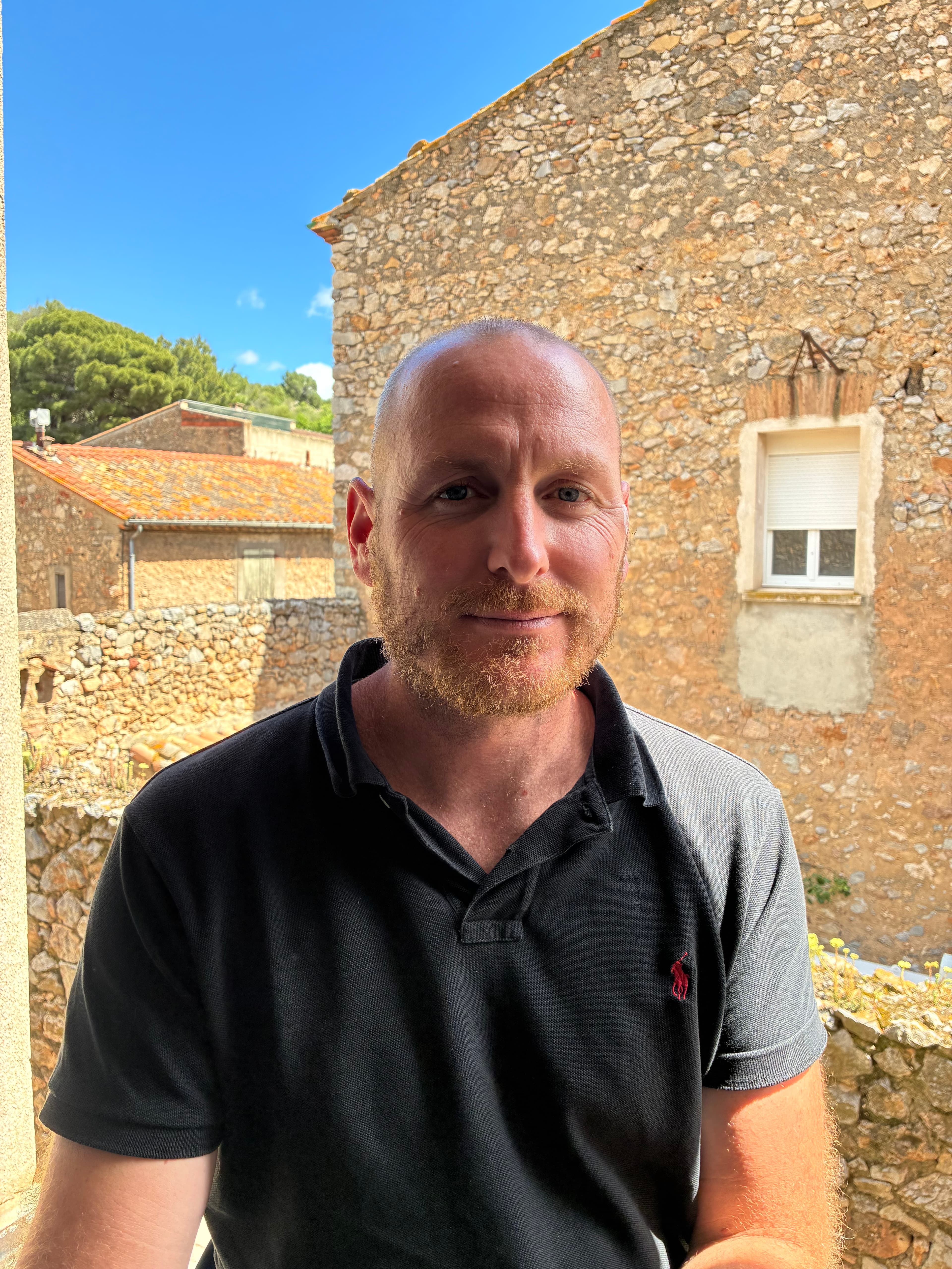 Homme à barbe rousse portant un polo noir devant une maison en pierre rustique.