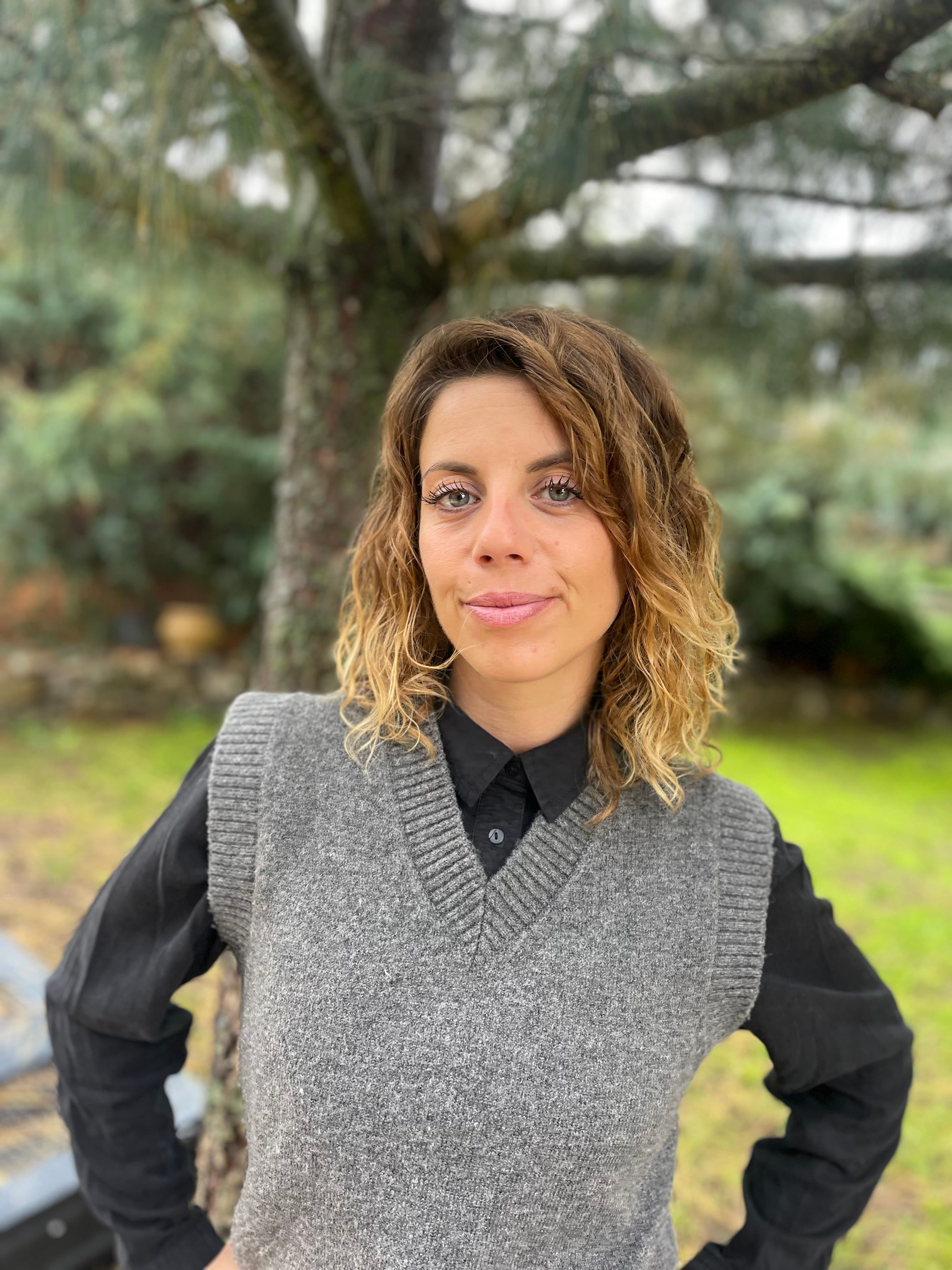 Femme aux cheveux ondulés portant un gilet gris sur une chemise noire en extérieur.